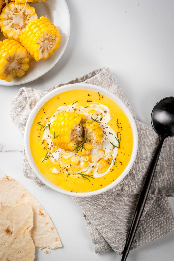 Vegan cuisine. Traditional autumn corn soup. Served with corn cobs, greens, cream. On a white marble table. Copy space top view