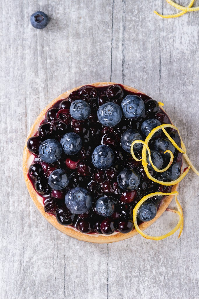 Round Lemon Tart with fresh and cooked blueberries, decorated by lemon zest over gray wooden surface. Top view