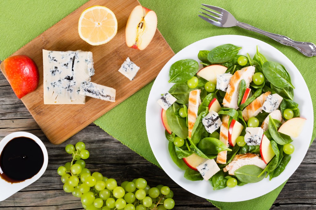 grilled chicken breast, grapes, spinach,  cheese and apple salad on a white dish, on a table mat, cutting board with pieces of apple, lemon and blue cheese, view from above