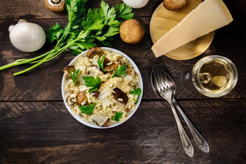 An overhead photo of a mushroom risotto, with champignons, fresh parsley, porcini, a slice of Parmesan, a glass of white wine, a fork and a spoon, and a place for text, on a dark rustic texture
